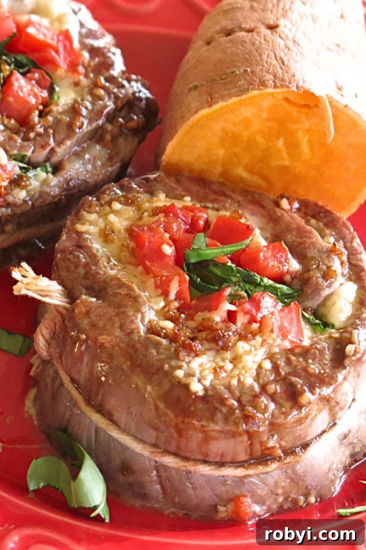 Flank steak pinwheel stuffed with spinach, mozzarella cheese, and tomatoes on a red plate with another pinwheel and a sweet potato behind it.