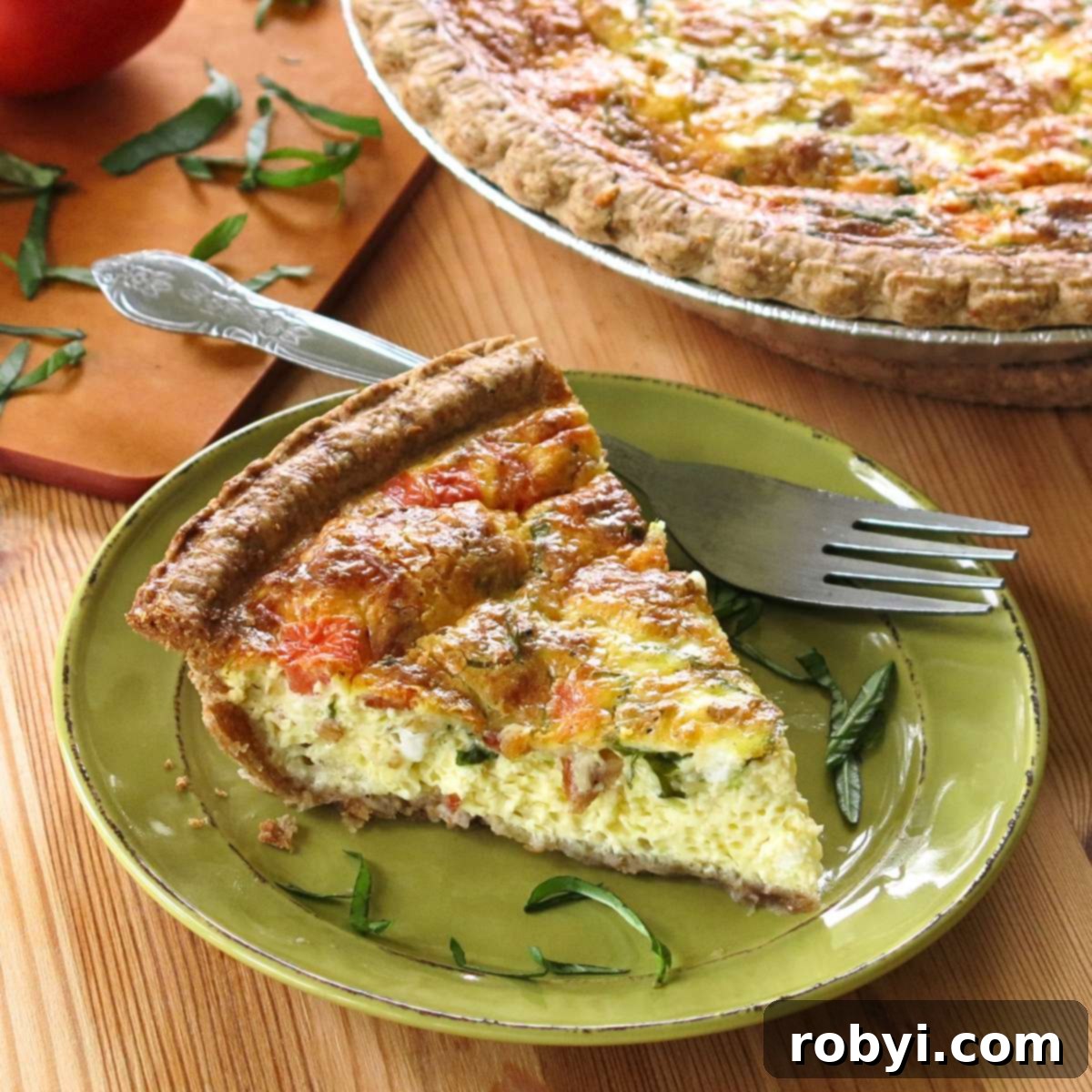 A perfectly baked slice of Tomato Bacon Quiche on a plate, with the rest of the golden-brown quiche in its pie dish in the background, showcasing its vibrant ingredients and flaky crust.