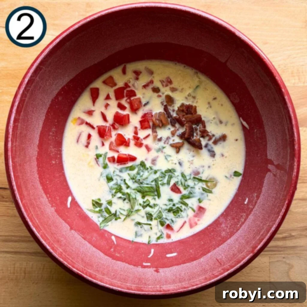 Diced tomatoes, crumbled bacon, and fresh chopped basil being carefully folded into the prepared egg and cheese mixture in a bowl, showing the vibrant colors.