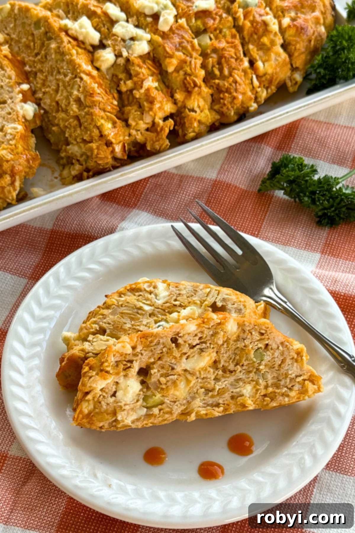 Two slices of buffalo chicken meatloaf on a plate with fork with a platter with more of the loaf behind it.
