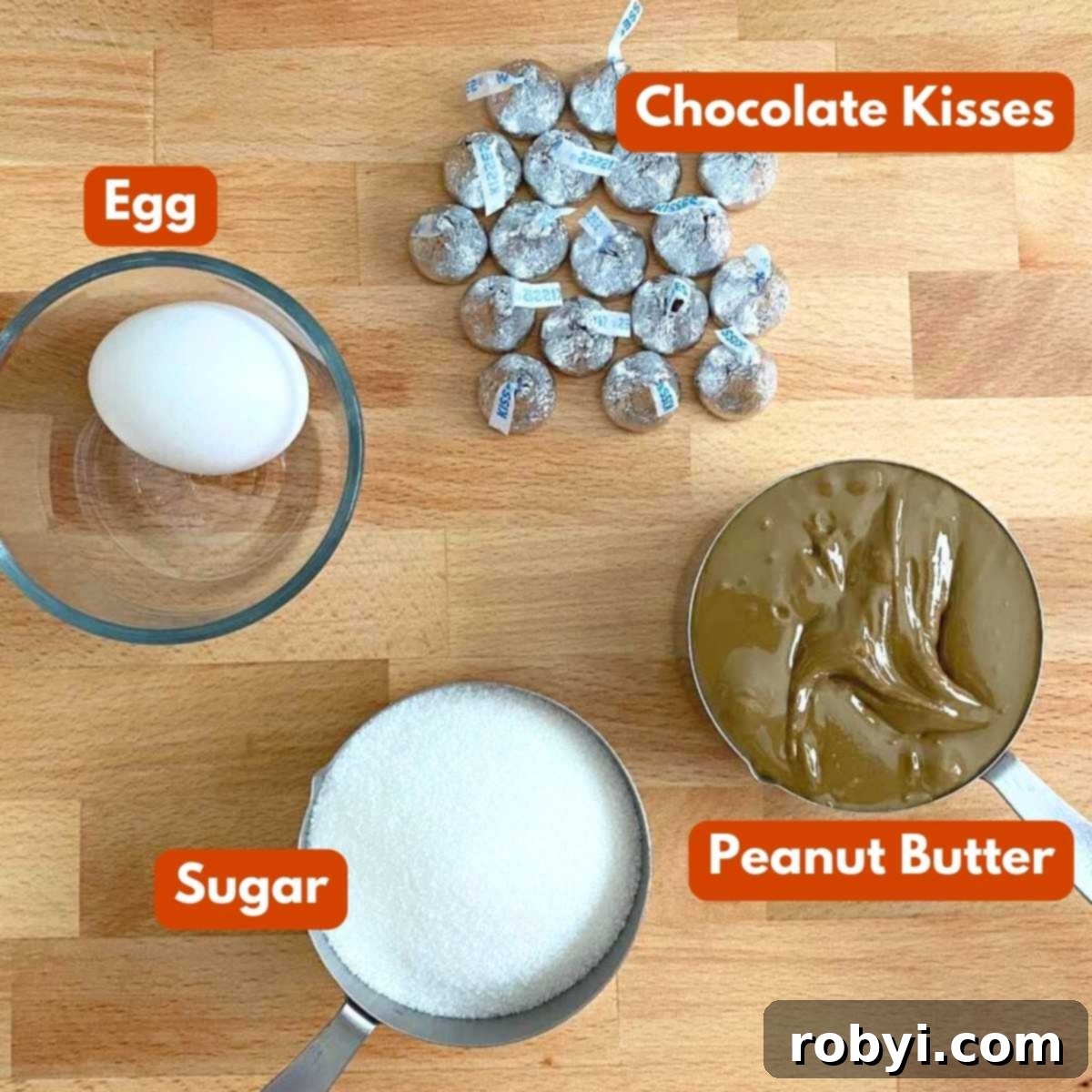A collection of ingredients laid out for making easy peanut butter blossom cookies: A jar of peanut butter, a bowl of sugar, a single egg, and several chocolate kisses.