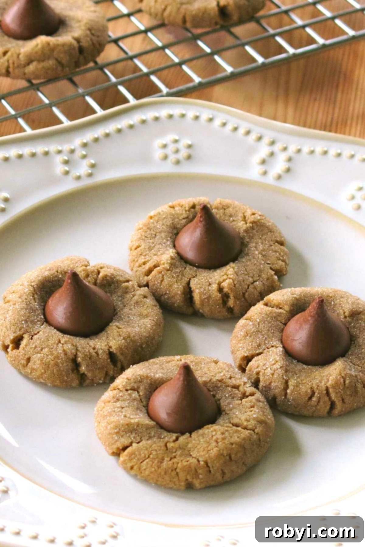 A charming display of three peanut butter blossoms on a small plate, with more cookies visible on a wire rack in the background.