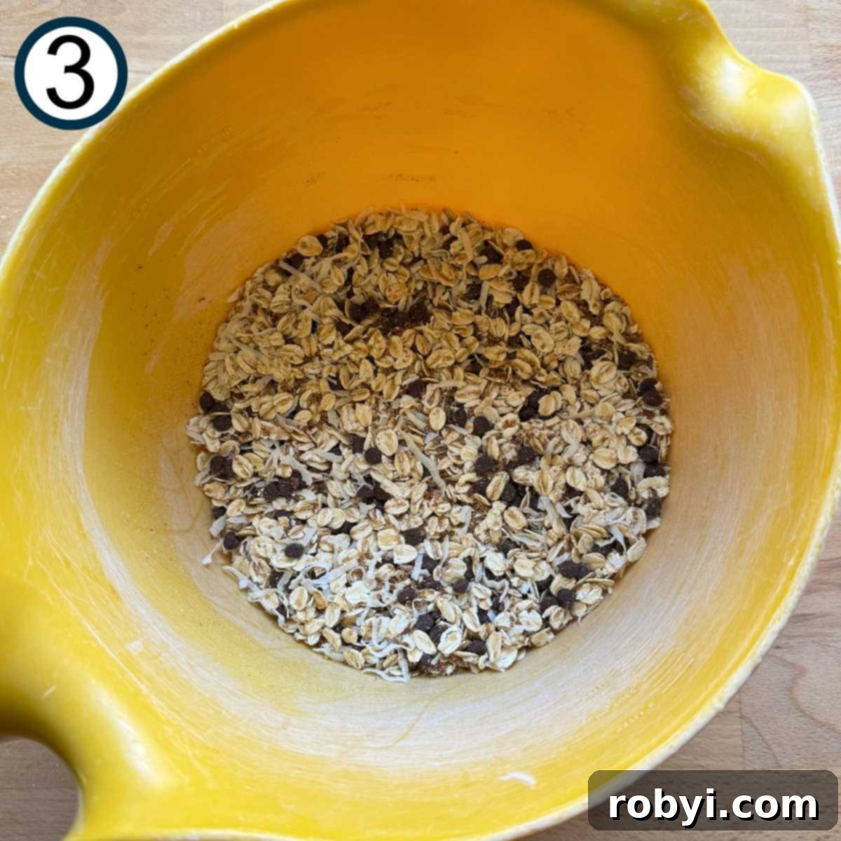 Oats, coconut flakes, ground flaxseed, and mini chocolate chips in a large yellow bowl.