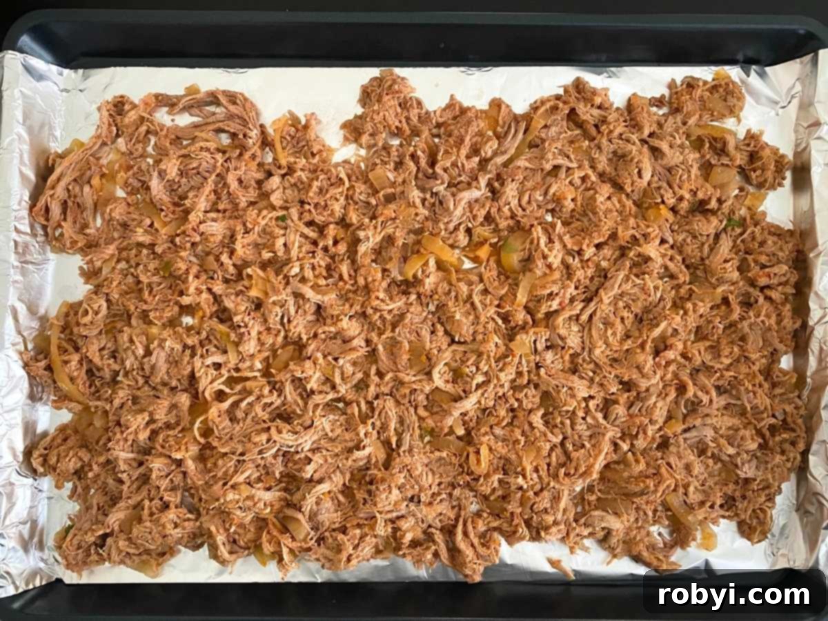 Shredded pork tenderloin carnitas spread out in an even layer on an aluminum foil-lined baking sheet, preparing for the crisping process under the broiler.
