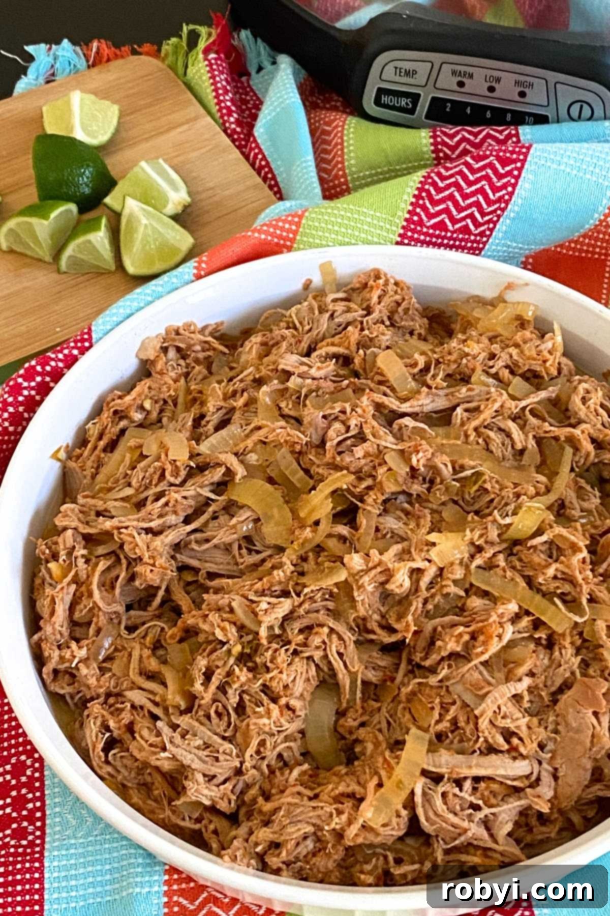 A large white serving dish filled with salsa pork tenderloin carnitas, accompanied by a slow cooker and fresh lime wedges in the background.