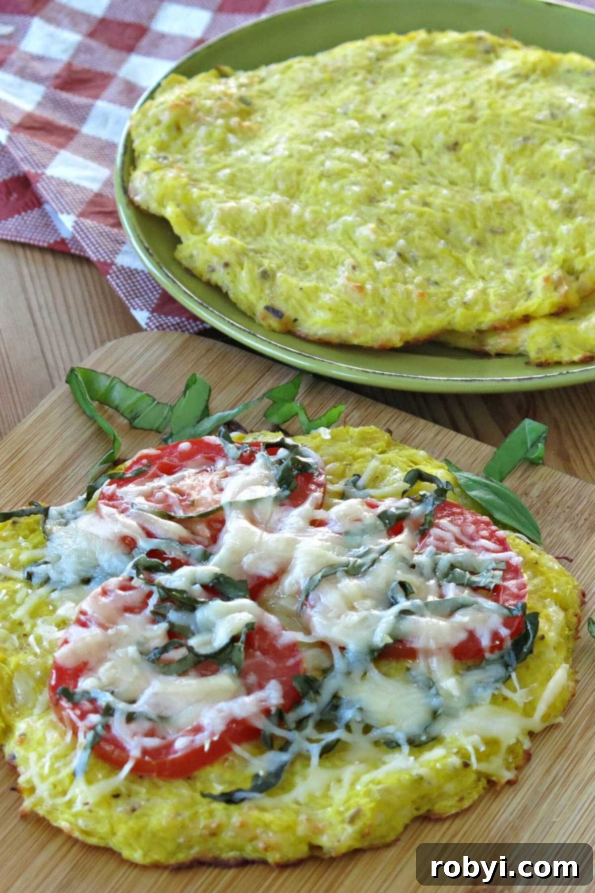 A vibrant spaghetti squash pizza crust adorned with fresh tomatoes, fragrant basil, and melted mozzarella cheese, positioned on a wooden cutting board with additional plain crusts stacked behind it.