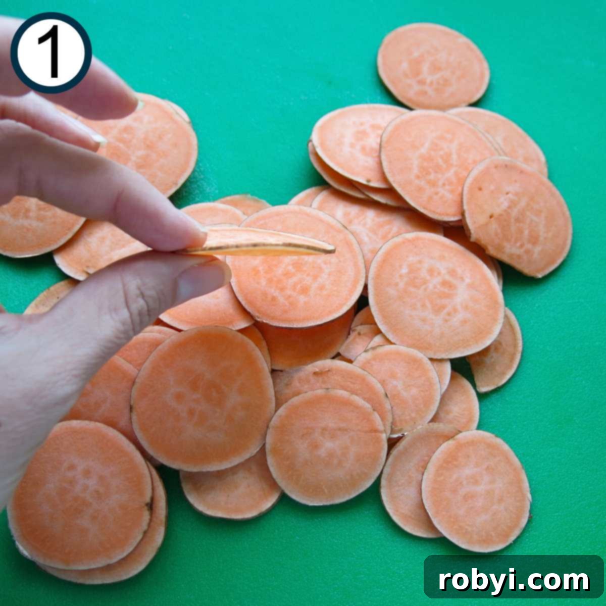 Fingers delicately holding a single, thinly sliced sweet potato disc above a mound of other uniformly sliced potatoes on a wooden cutting board.