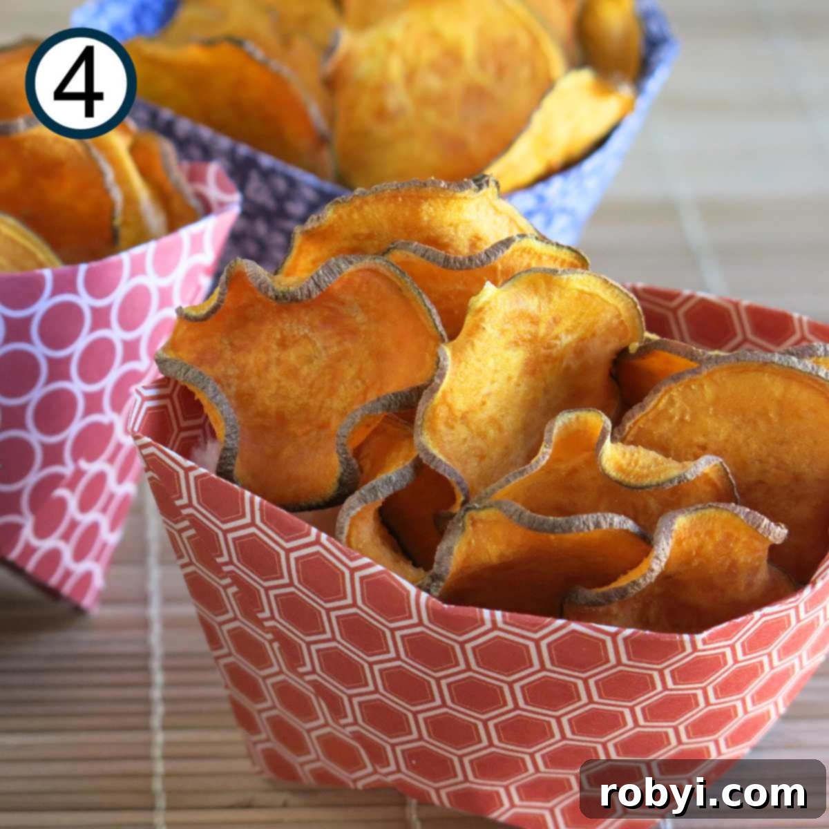 A collection of perfectly crispy sweet potato chips, with appealingly curled edges, presented in a neat paper container, with more containers blurred in the background.