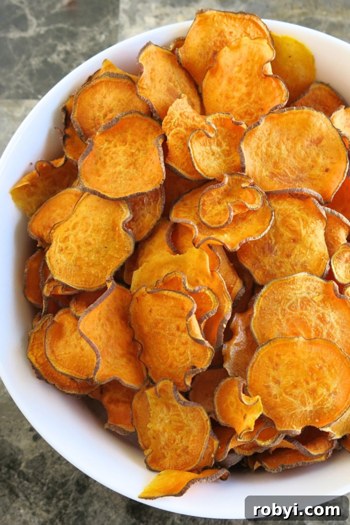 A generously sized white bowl overflowing with a beautiful mound of freshly oven-baked sweet potato chips, set against an elegant marble background, highlighting their rustic appeal and golden crispness.