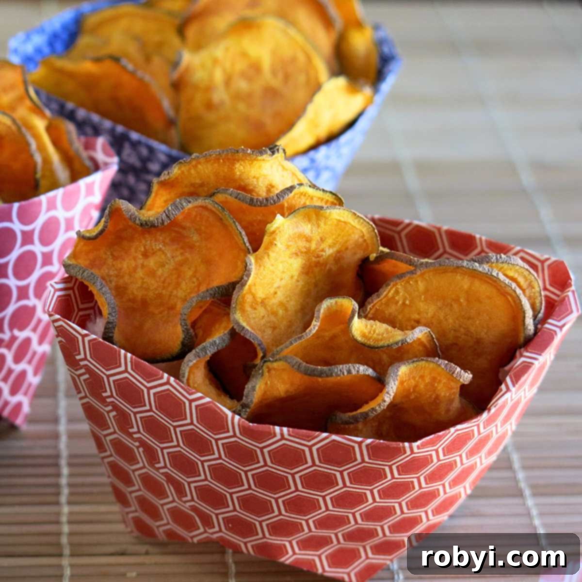 A charming close-up of baked sweet potato chips nestled in a paper container, with two more containers filled with chips subtly blurred in the background.