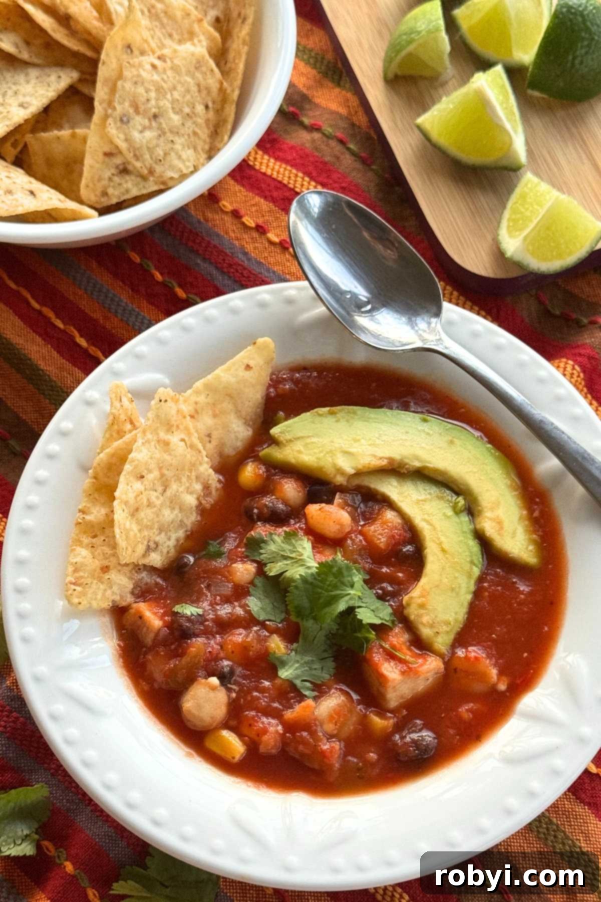 Serving bowl and spoon with chicken tortilla soup showing hominy, black beans, and corn. It's topped with cilantro, 2 slices of avocado, with 3 tortilla chips are tucked on the side. A bowl of tortilla chips and lime wedges are behind it.