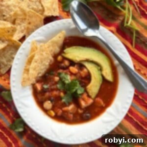 Serving bowl and spoon with chicken tortilla soup showing hominy, black beans, and corn. It's topped with cilantro, 2 slices of avocado, with 3 tortilla chips are tucked on the side.