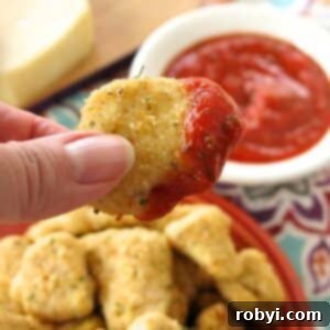 Fingers holding a garlic parmesan chicken bite that has been dipped in marinara sauce over a plate with more.