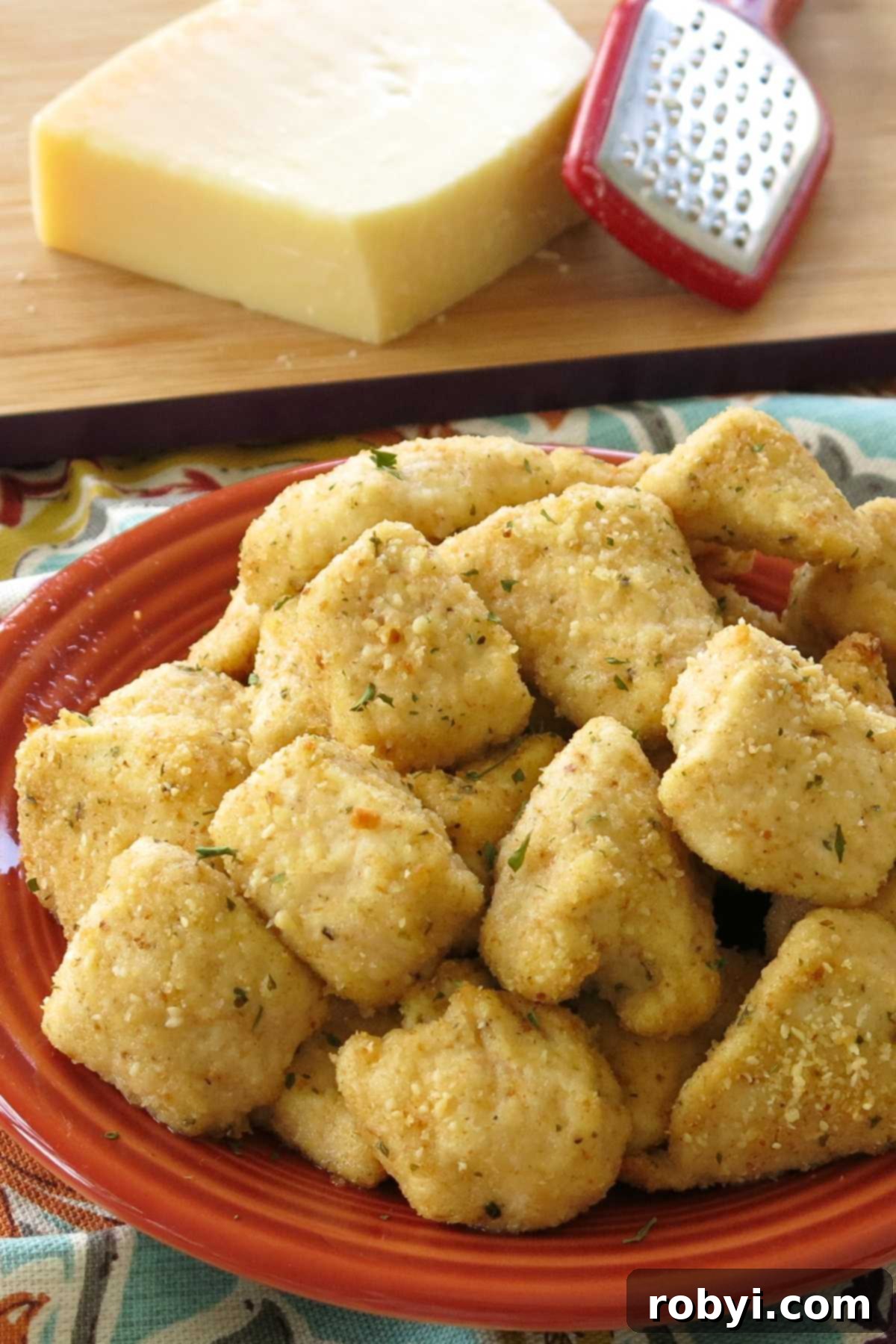A generous pile of golden-brown garlic parmesan chicken bites on a white plate, with a block of Parmesan cheese and a grater artfully placed behind it.