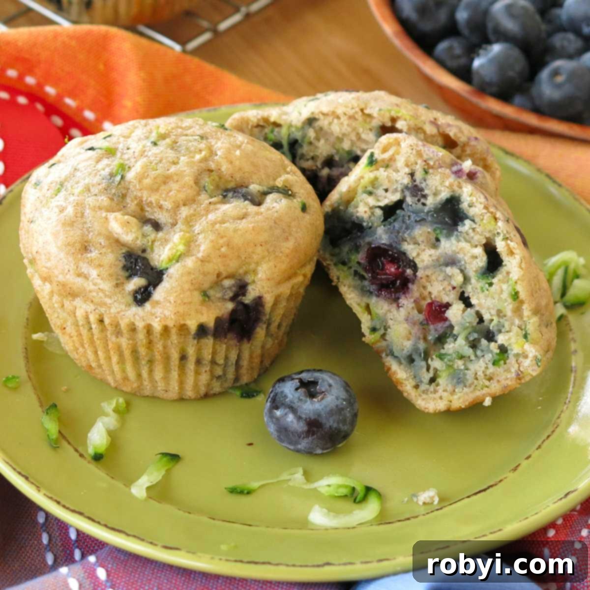 Two zucchini blueberry muffins made with Greek yogurt on a plate, with one cut half with fresh blueberries behind it and a few shreds of zucchini on the plate.