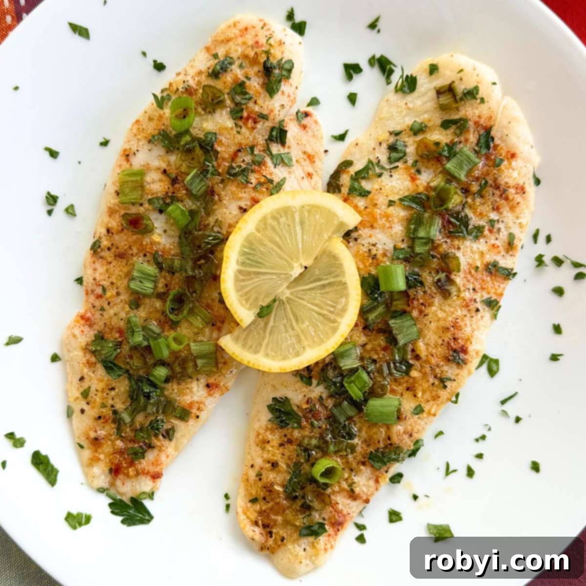 Two lemon baked basa fillets topped with lemon slices on a white plate that is scattered with parsley. A healthy, zesty, oven-baked white fish meal, ready in 25 minutes.
