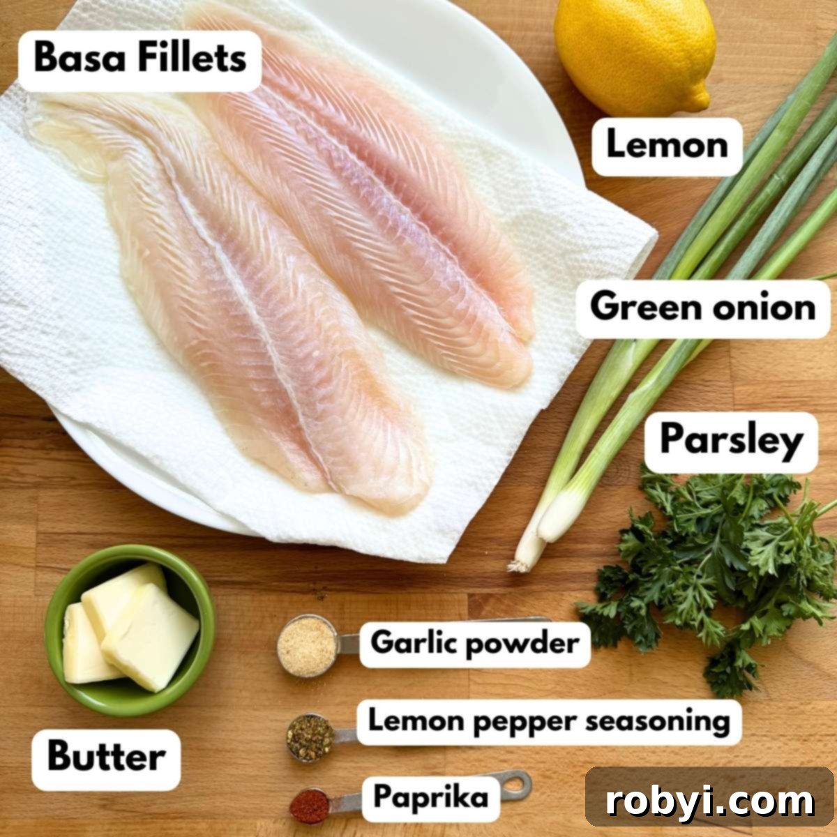 Two basa fillets, a lemon, two green onion, tabs of butter, parsley, and garlic powder, lemon pepper seasoning, and paprika on a cutting board, ready for baking.