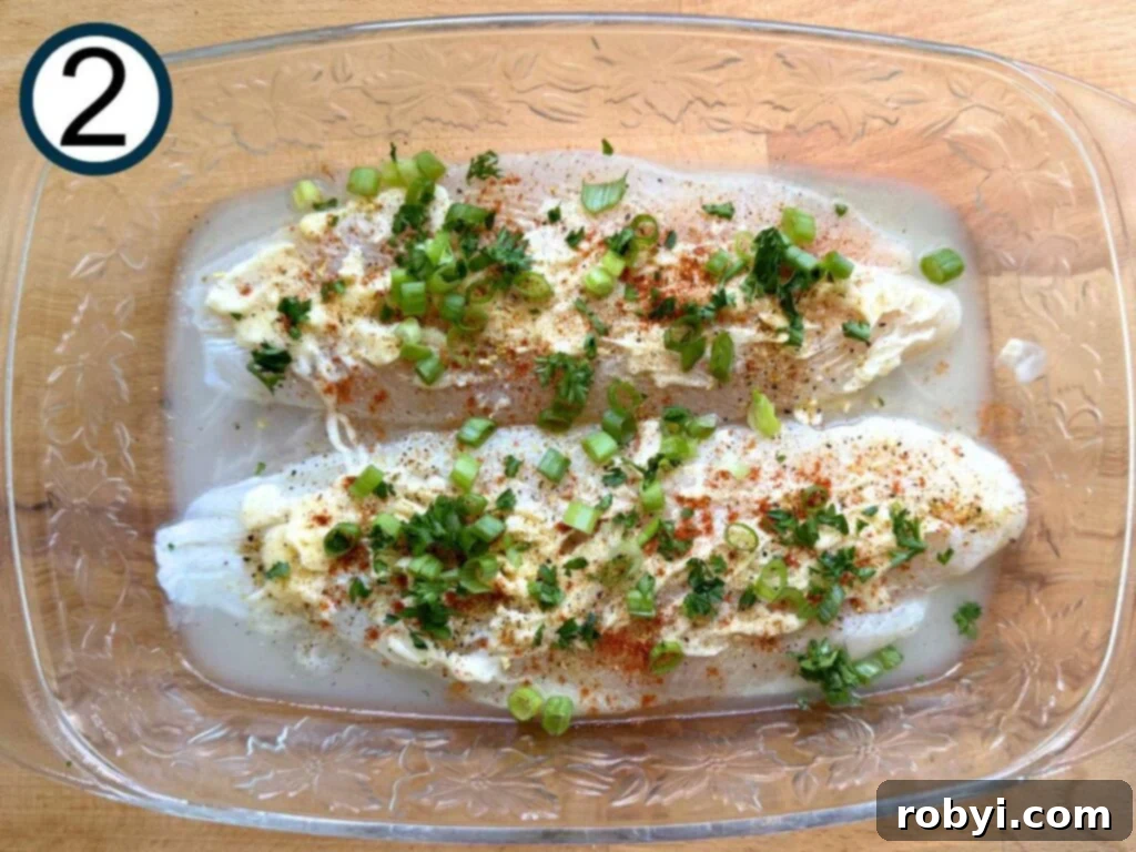 Two uncooked basa fillets topped with a generous layer of spices, chopped green onion, and fresh parsley, in a clear glass baking dish.