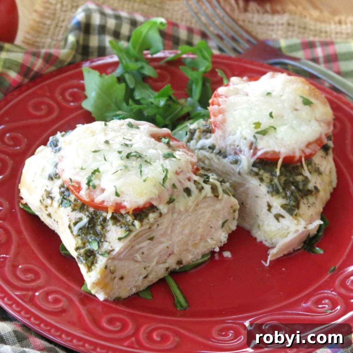Baked pesto chicken with tomatoes and mozzarella cheese cut in half on on a red plate with arugula and a fork in background.