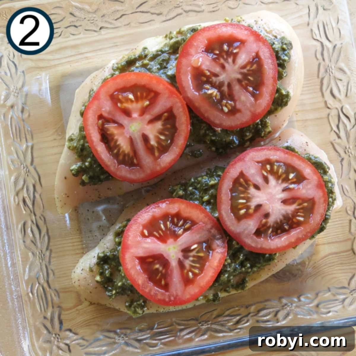 Slices of tomato on top of two boneless, skinless chicken breasts with pesto spread on top in a glass baking dish.