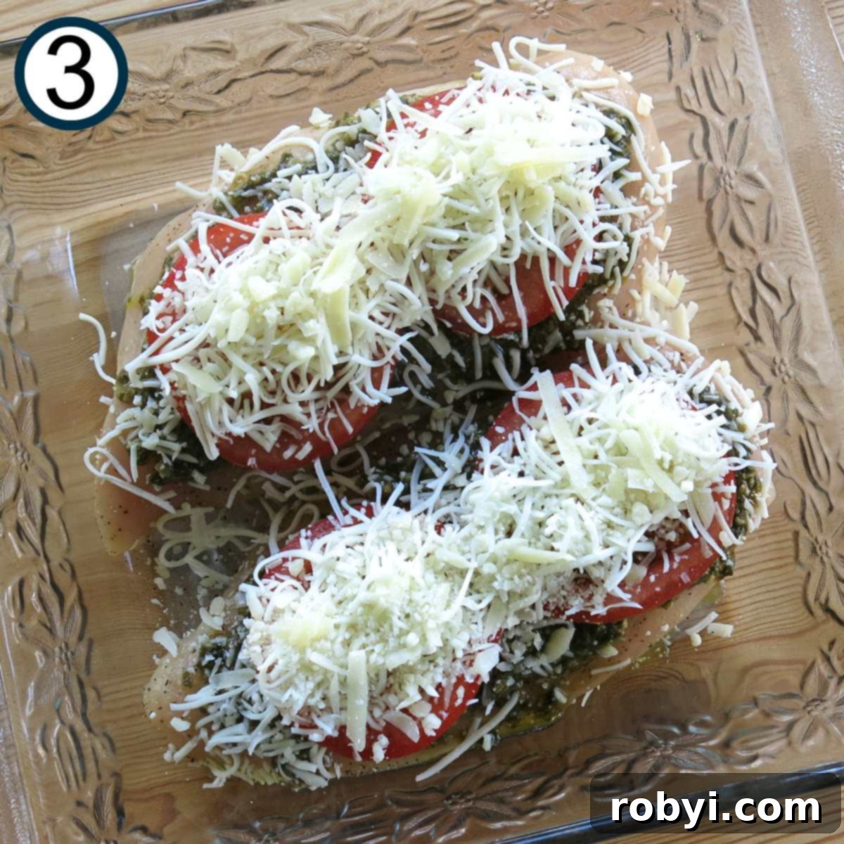 Shredded mozzarella cheese layered on top of two chicken breasts layered with pesto and tomatoes in a glass baking dish.