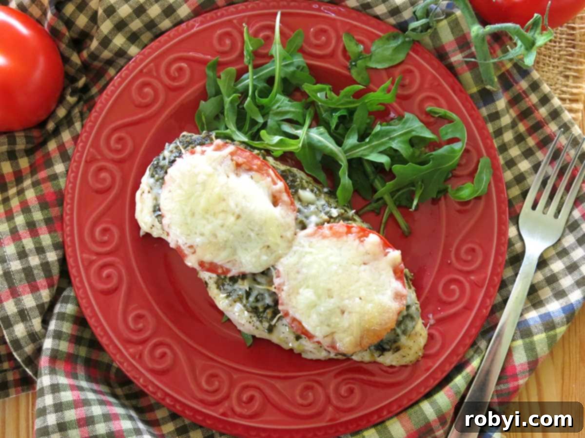 Baked chicken breast topped with pesto, sliced tomatoes, mozzarella, and Parmesan cheese on a red plate with a little arugula.