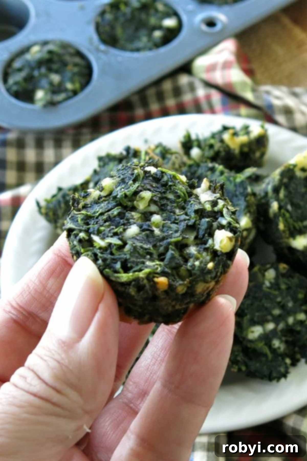 A hand gently holds one perfectly baked keto spinach feta egg cup, with a full pan of additional egg muffins blurring in the background, ready for enjoyment.
