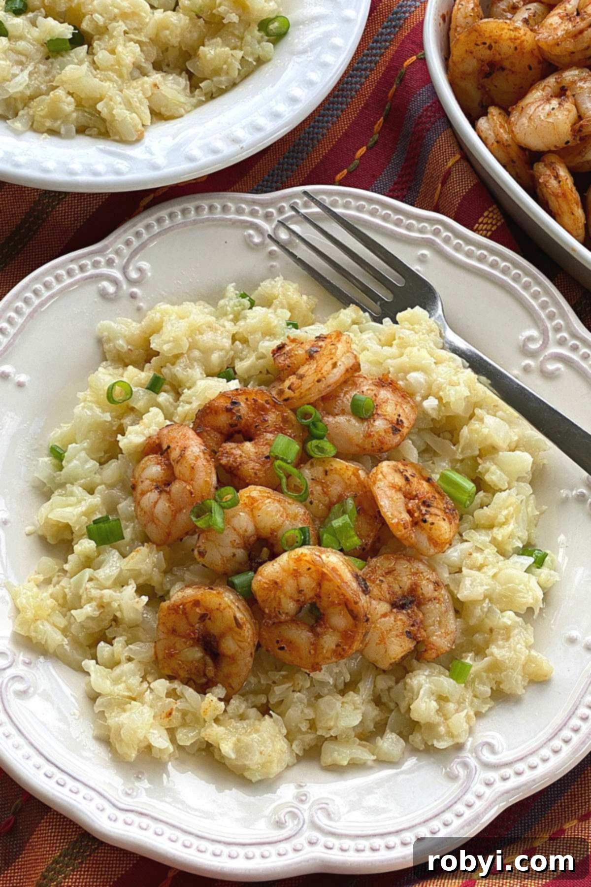Blackened shrimp on top of a plate with cauiflower grits with more shrimp and a bowl of grits in the background.