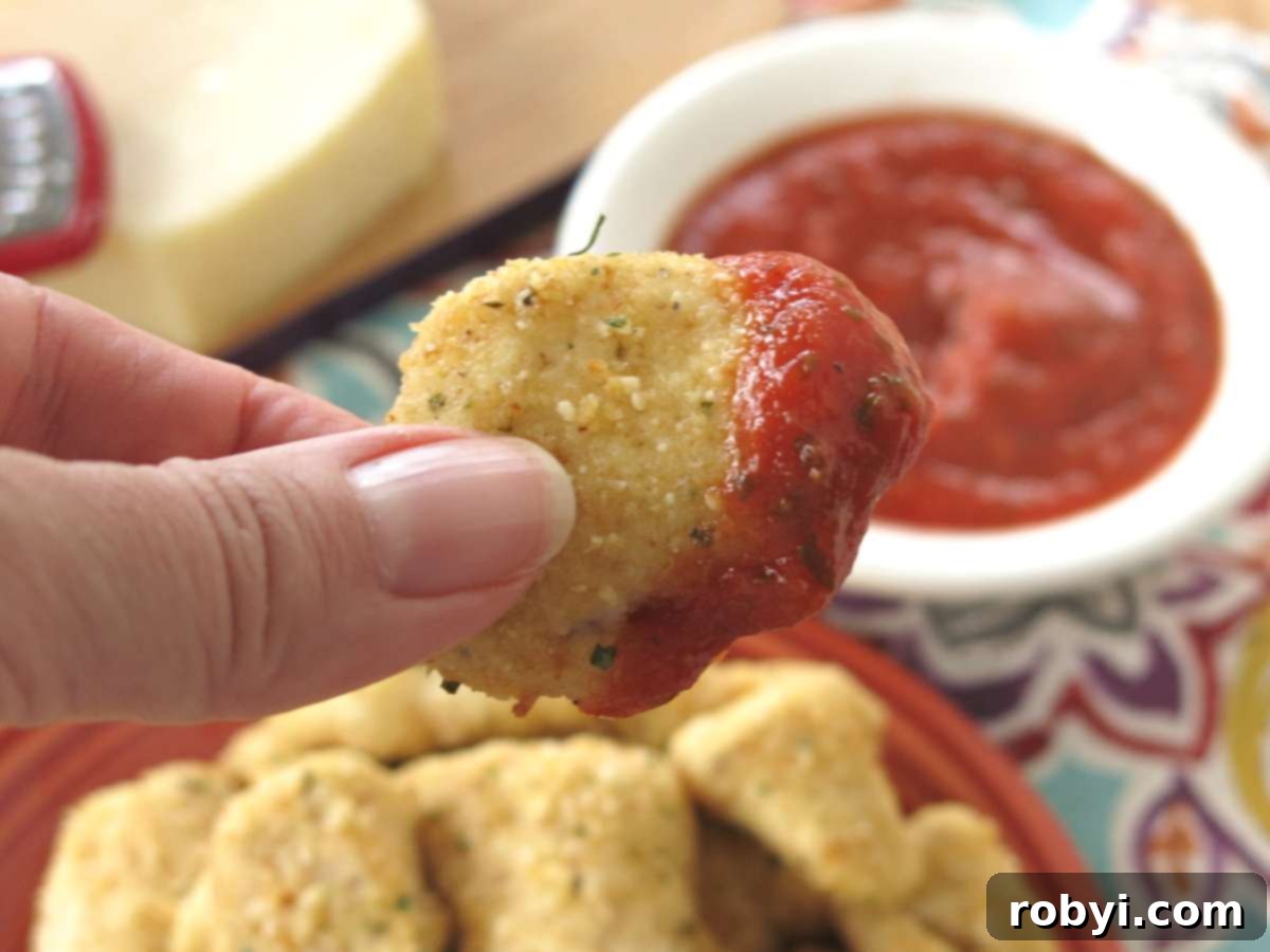 Fingers holding a Parmesan chicken nugget dipped in marinara over a plate with more nuggets.