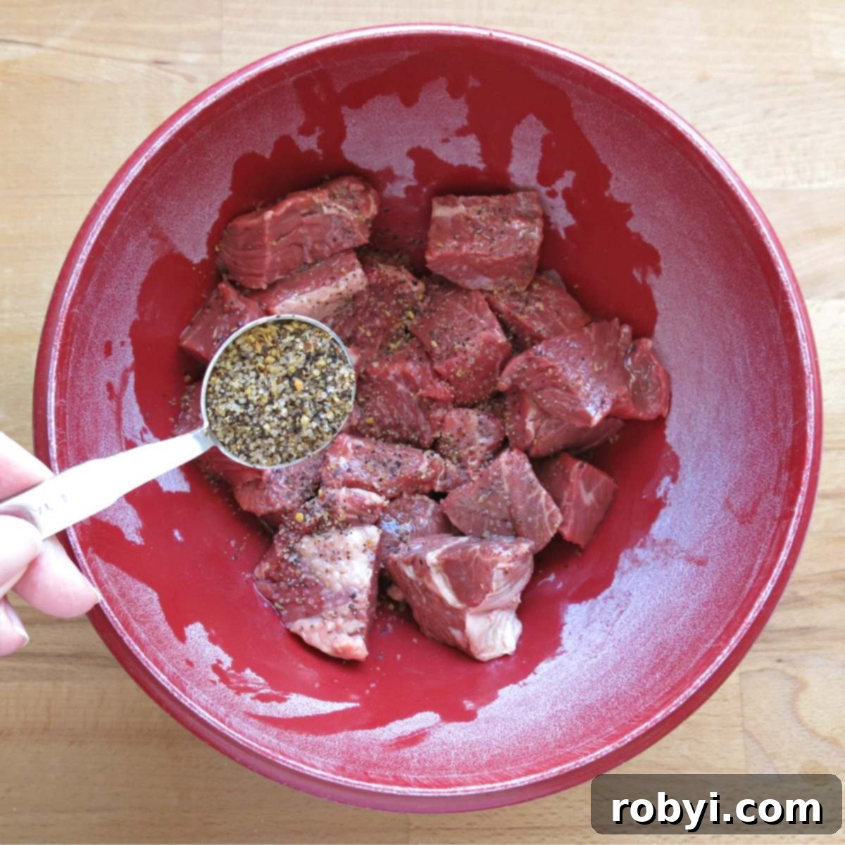 Steak seasoning in a measuring spoon being sprinkled over cubed beef in a vibrant red bowl.