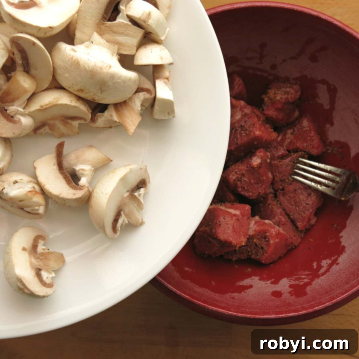 Chopped mushrooms being added from a plate into a bowl containing seasoned beef cubes.
