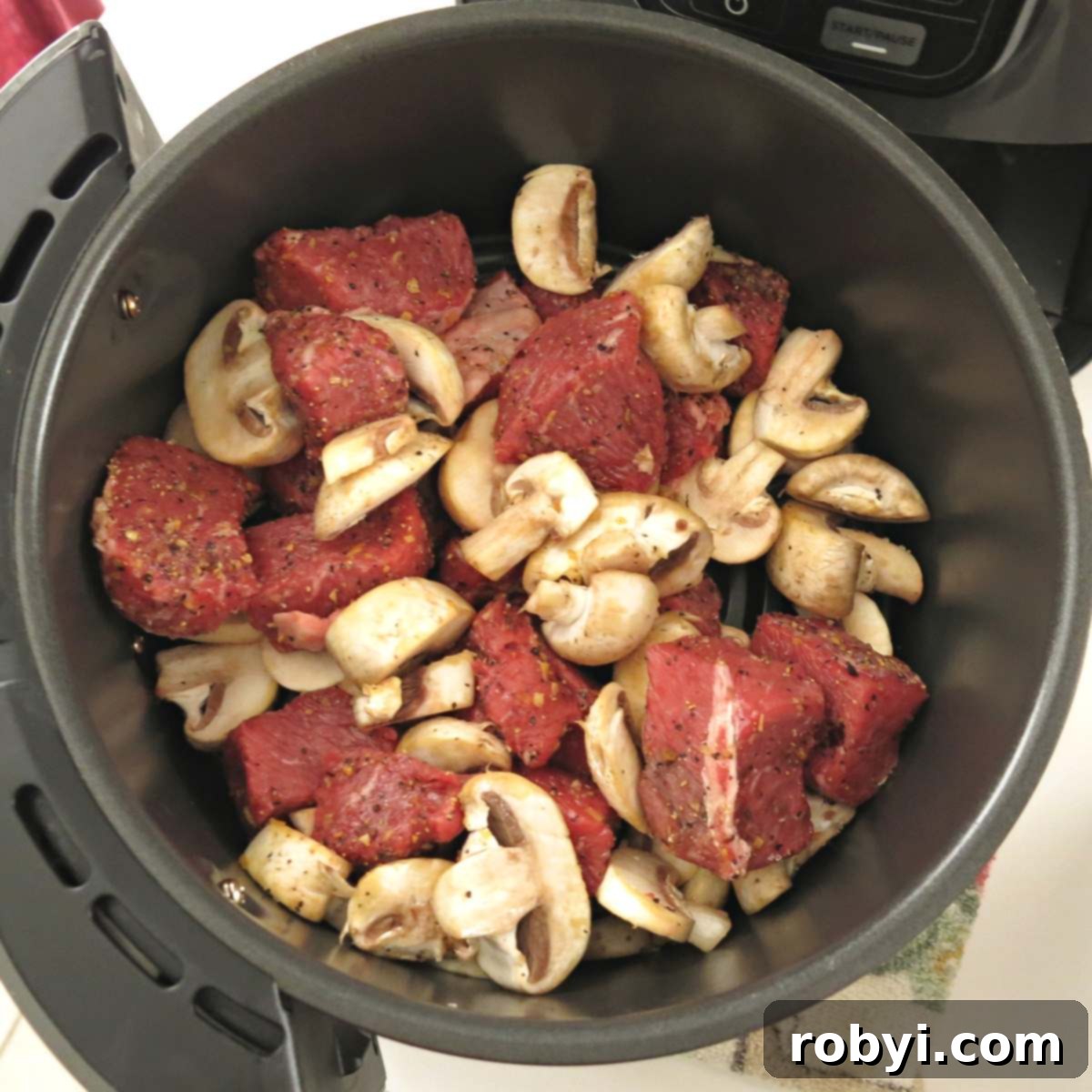 Uncooked sirloin steak cubes and sliced mushrooms neatly arranged in an air fryer basket.