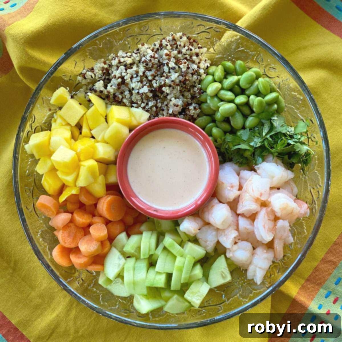 All the fresh ingredients for a Shrimp Poke Bowl laid out before combining: shrimp, rice, edamame, carrots, cucumbers, mango, cilantro, and a bowl of creamy poke sauce in the center.