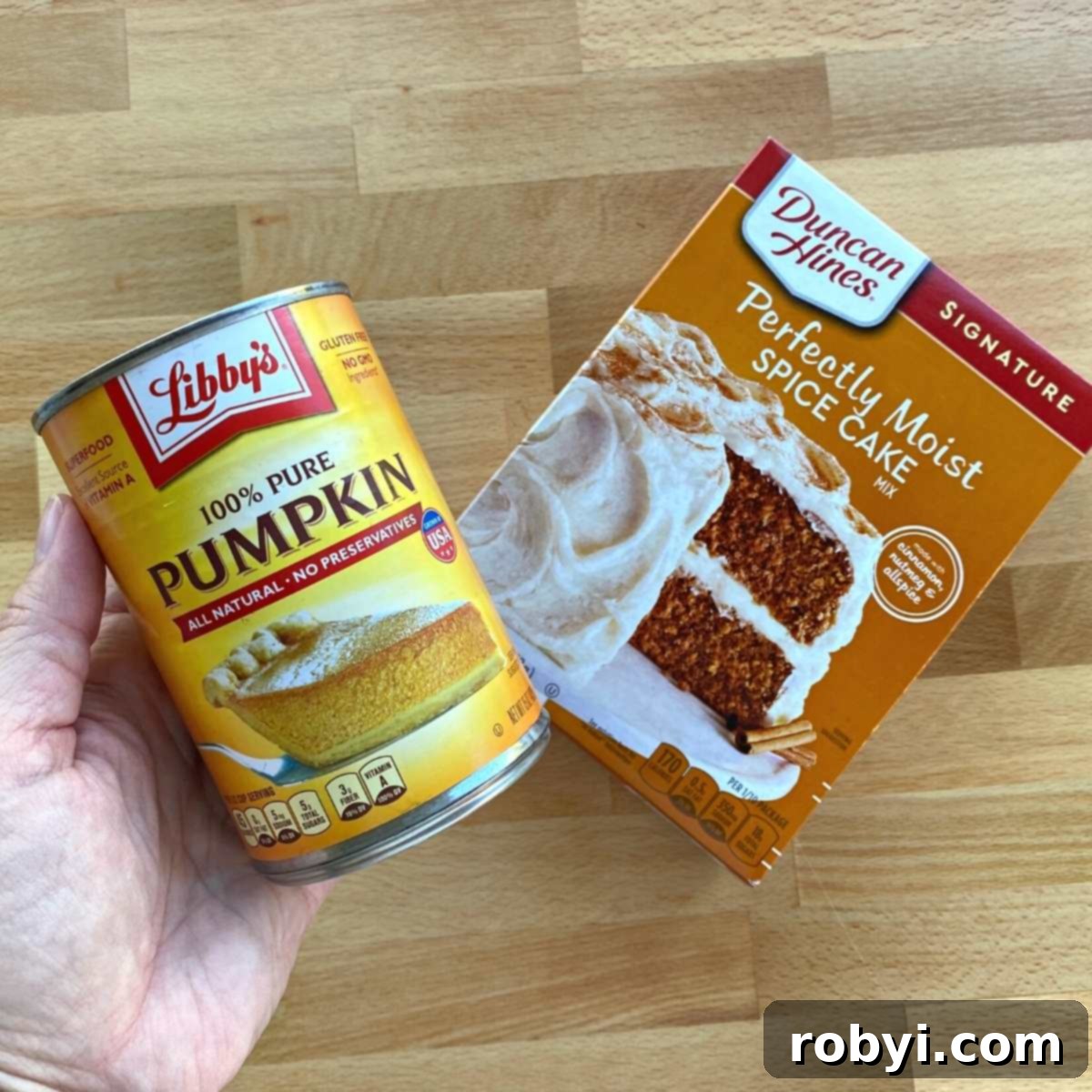 A hand holding a can of pumpkin puree next to a box of Duncan Hines Spice Cake mix, highlighting the two simple ingredients.