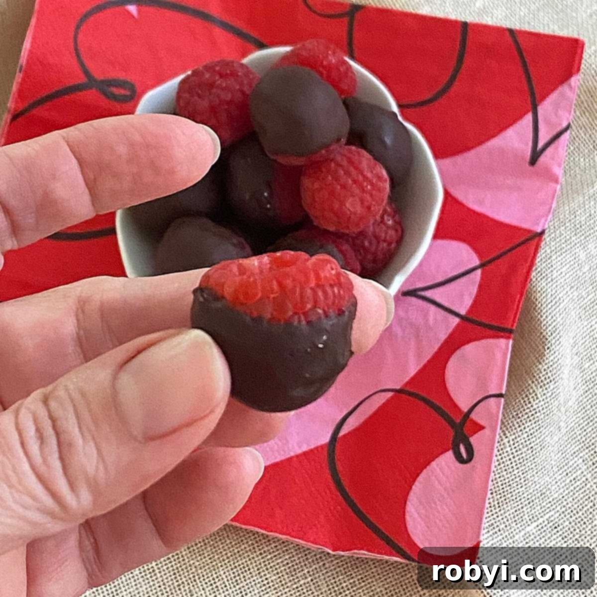 Chocolate-Dipped Frozen Raspberry Jewels 5 A hand delicately holding a single dark chocolate dipped raspberry, highlighting the perfect coating and inviting texture of the frozen treat.