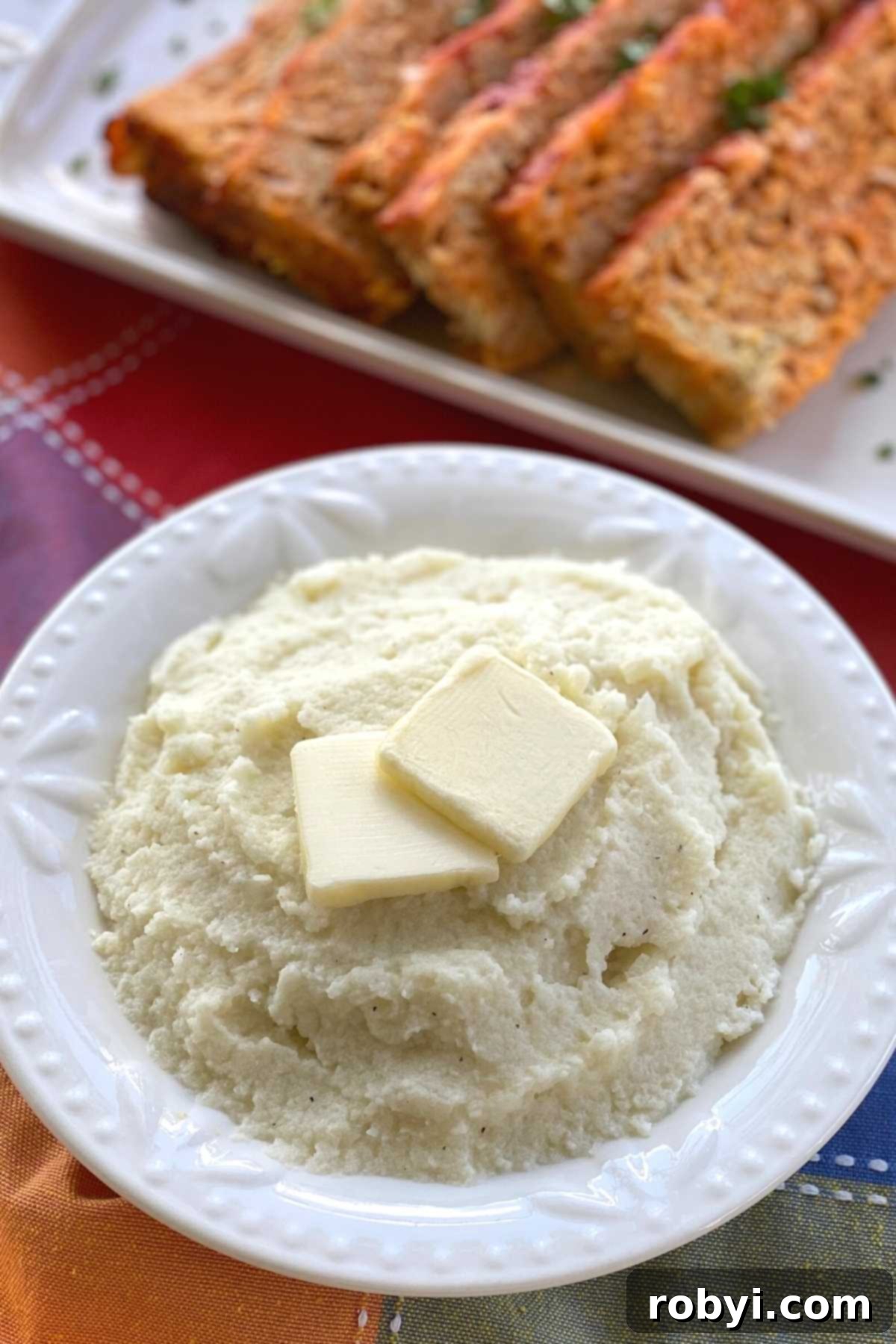 Creamy Cauliflower Mashed Potatoes in a bowl next to a platter of savory meatloaf.