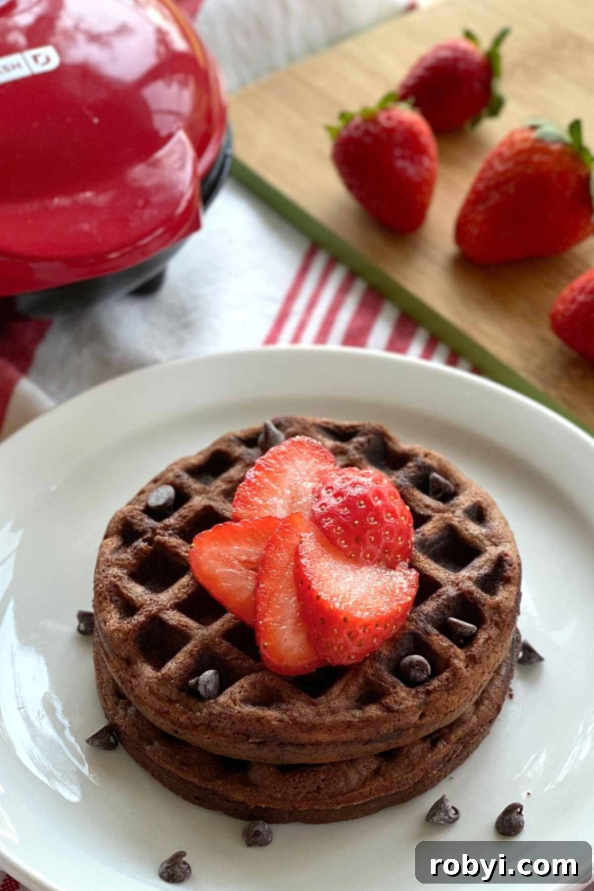 Decadent Chocolate Chaffles with Ricotta 5 A plate featuring two chocolate waffles, accompanied by a waffle iron and fresh strawberries in the background.