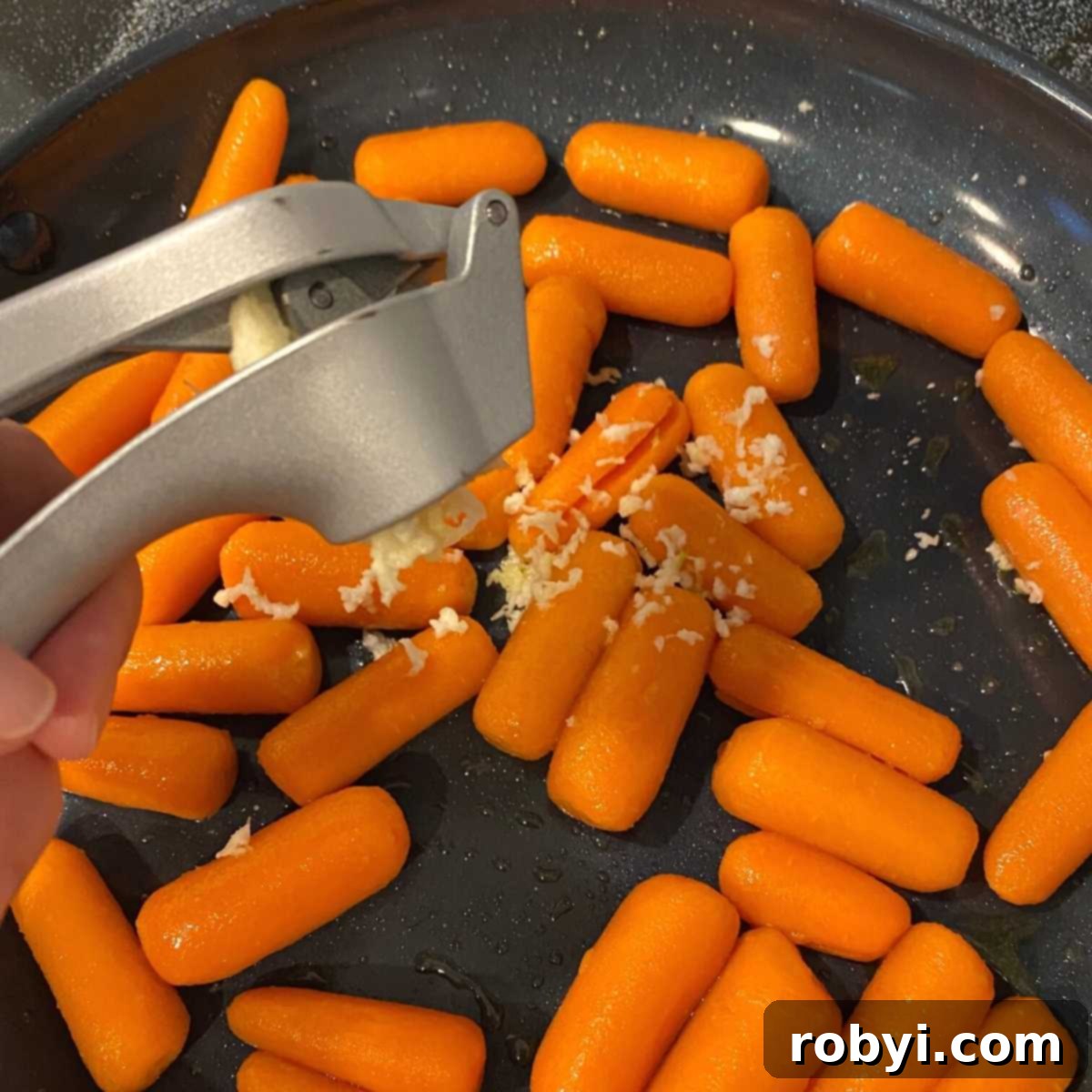 Garlic being added to a skillet of baby carrots with garlic press.