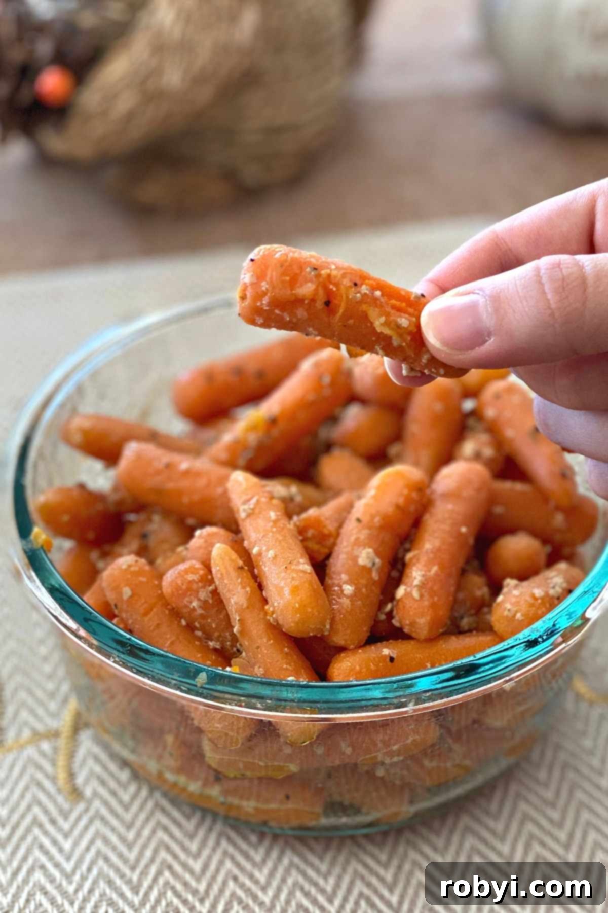 Fingers holding a sauteed baby carrot with a glass bowl with more below.
