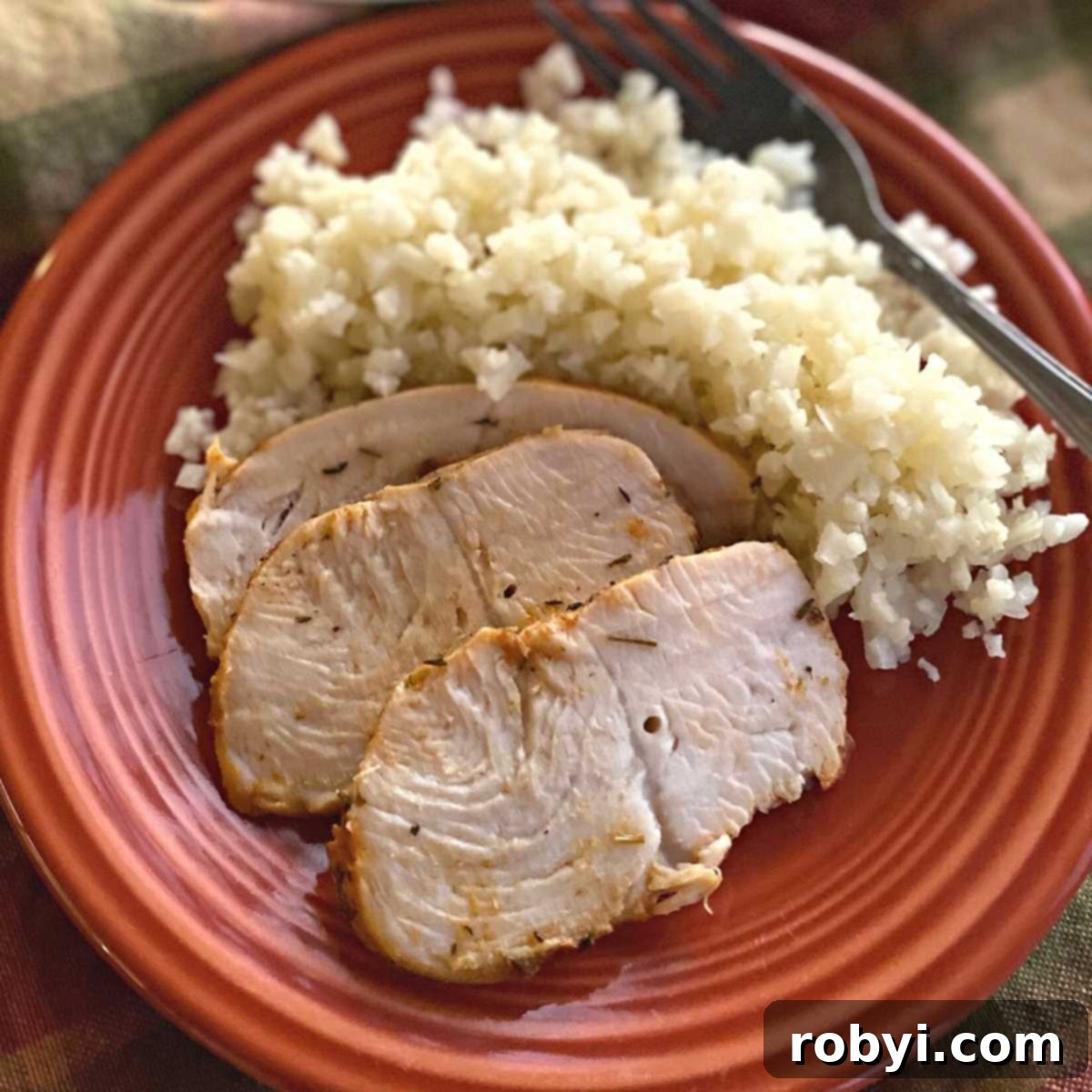 Three perfectly sliced pieces of baked turkey tenderloin served on a white plate alongside fluffy cauliflower rice and a fork, highlighting its succulent texture.