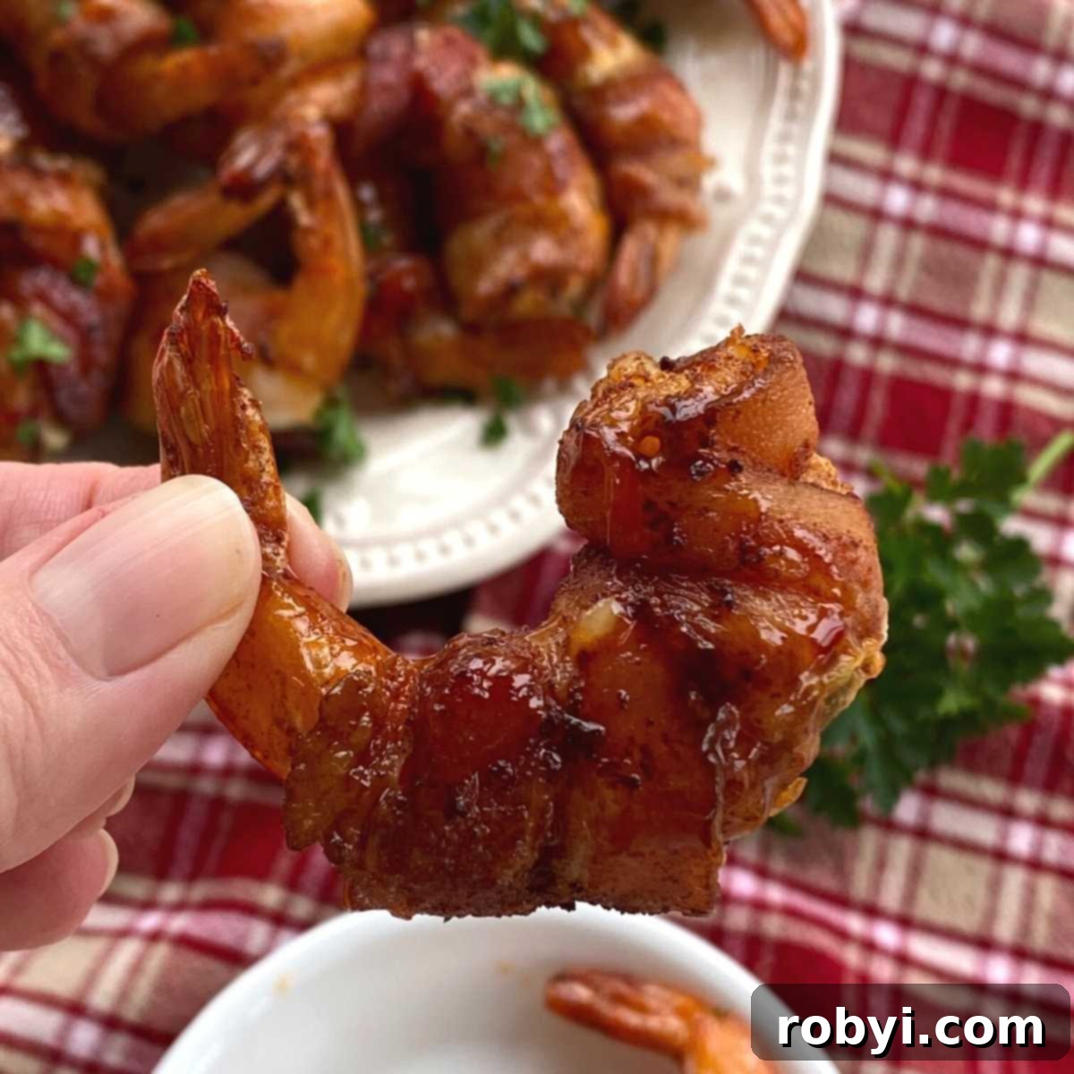 Fingers holding a perfectly cooked stuffed bacon wrapped shrimp, with more beautifully arranged on a plate in the background.