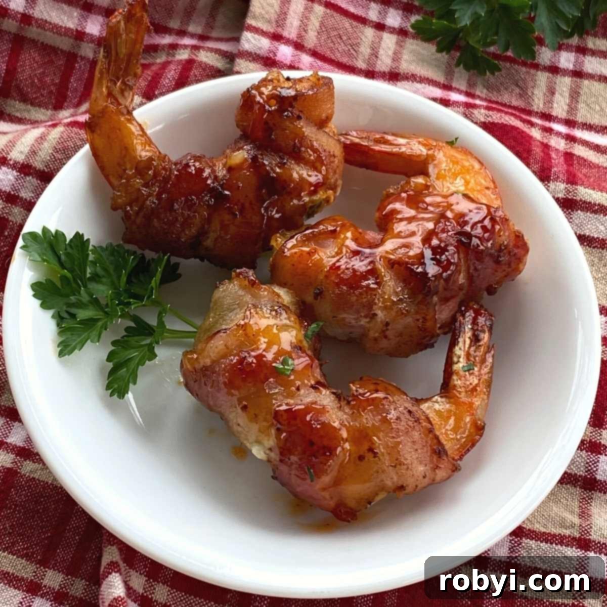Three perfectly golden-brown bacon wrapped shrimp, stuffed with cream cheese and a jalapeno slice, artfully arranged on a white plate.