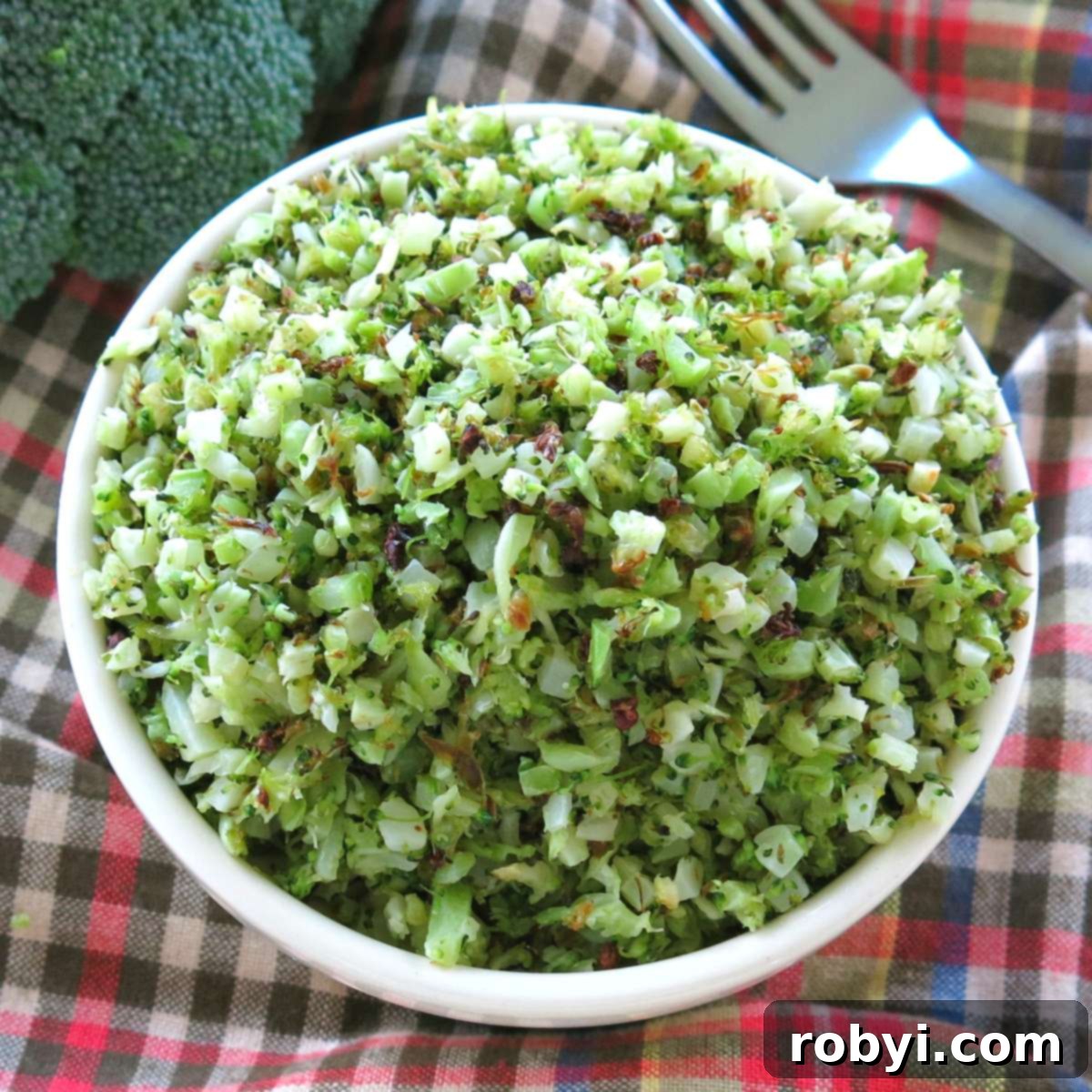 A bowl of roasted riced broccoli, next to a fresh head of broccoli and a fork.