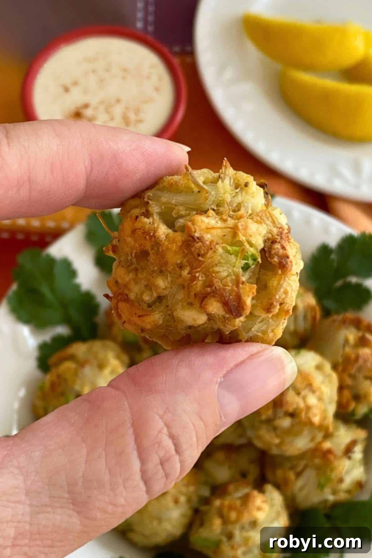 Fingers holding a crab ball appetizer over a bowl with more.