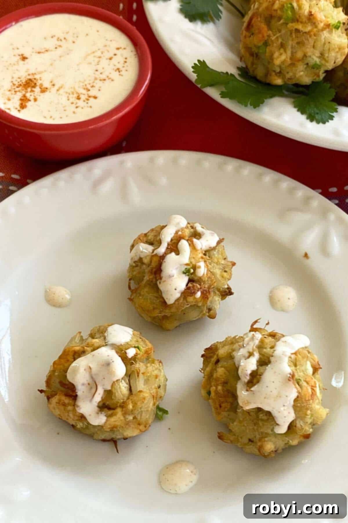 Three crab bites drizzled with aioli on a plate, garnished with fresh herbs.
