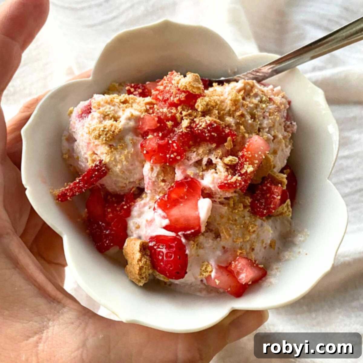 Hand holding a tulip-shaped cup with strawberry cottage cheese ice cream topped with strawberries and graham crackers.