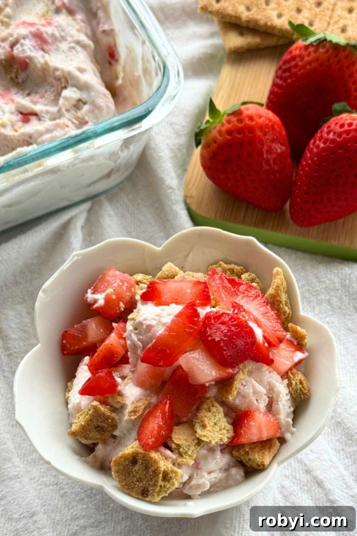 Strawberry cheesecake cottage cheese ice cream in a tulip bowl with fresh strawberries, graham crackers, and the container with more ice cream behind it.
