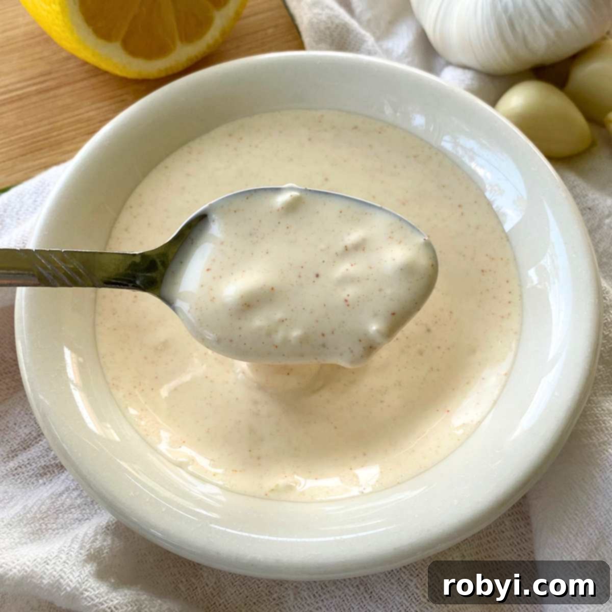 Homemade Lemon Garlic Aioli: Simple & Delicious 5 A silver spoon gracefully lifts a generous scoop of creamy homemade lemon garlic aioli from a small, elegant white bowl, showcasing its rich texture and inviting appearance, ready to be enjoyed.