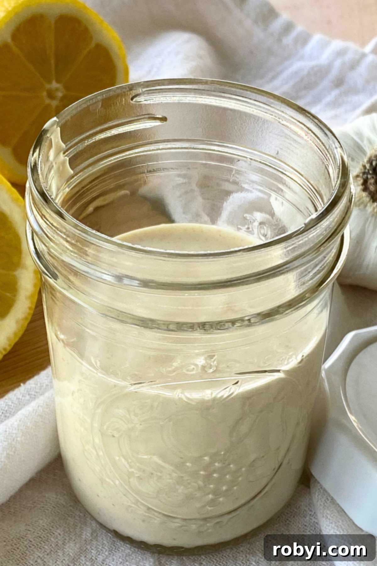 Homemade Lemon Garlic Aioli: Simple & Delicious 6 A small mason jar filled with easy lemon garlic aioli, tightly sealed with a lid, sits on a rustic wooden surface. Beside it, a fresh whole lemon and a head of garlic reinforce the natural, wholesome ingredients used in the sauce.