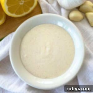 Homemade Lemon Garlic Aioli: Simple & Delicious 7 Lemon garlic aioli sauce in a bowl with a cut lemon and garlic cloves behind it.